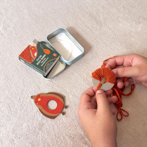 Cotton Twist Rosy Robin pom pom decoration kit inside an open metal tin, featuring a wooden robin cutout and yarn.